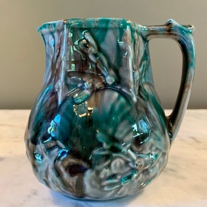 May include: A green and brown ceramic pitcher with a textured surface and a floral design. The pitcher has a handle and a wide mouth.