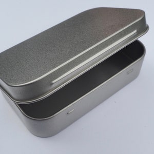 Peut inclure: Une boîte rectangulaire en métal argenté avec un couvercle à charnière. La boîte est ouverte et le couvercle repose sur le dessus de la boîte.