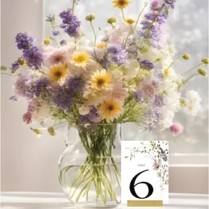 Op de afbeelding: Een bloemige tafelnummerkaart met een witte achtergrond en een gouden rand. De kaart heeft een aquarel-illustratie van paarse en roze bloemen en groen. Het nummer "6" is in zwart op de kaart gedrukt. De kaart ligt op een tafel voor een vaas met bloemen.