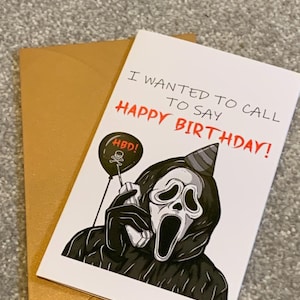 May include: A birthday card featuring a Ghostface character illustration, holding a phone and wearing a party hat. The card has the text "I WANTED TO CALL TO SAY HAPPY BIRTHDAY!" in red. A gold envelope is next to the card.