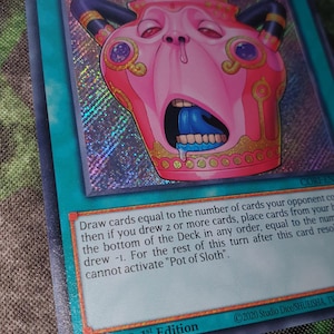 May include: A holographic trading card titled Pot of Sloth, a spell card with a teal border. The central illustration features a pink, anthropomorphic pot with a wide-open mouth, blue tongue, and dark horns, set against a shimmering, rainbow-colored background.