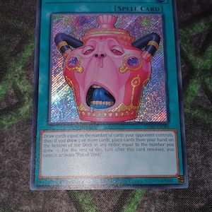 May include: A holographic Yu-Gi-Oh! trading card titled Pot of Sloth, a Spell Card. The card features a pink, pot-like creature with a wide-open mouth, blue tongue, and dark horns. The text box details the card effect, and the card code is CORI-EN061.