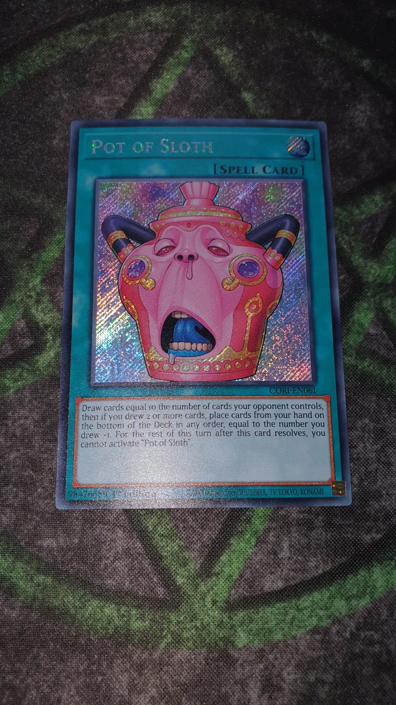 May include: A holographic Yu-Gi-Oh! trading card titled Pot of Sloth, a Spell Card. The card features a pink, pot-like creature with a wide-open mouth, blue tongue, and dark horns. The text box details the card effect, and the card code is CORI-EN061.