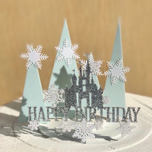 Könnte beinhalten: Ein weißer Kuchen, verziert mit einem silbernen Glitzer-Schloss, blauen und weißen Bäumen und weißen Schneeflocken. Der Kuchen hat außerdem einen silbernen Glitzer-„Happy Birthday“-Topper.
