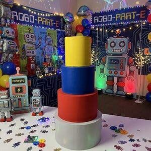 Puede incluir: Una tarta de varios niveles con capas amarillas, azules, rojas y blancas es la pieza central de una fiesta temática de robots. Las decoraciones de la fiesta incluyen globos de robots, pancartas que dicen "ROBO-PART-E" y figuras de robots. Confeti con forma de robot y decoraciones de placas de circuito están esparcidos sobre la mesa.