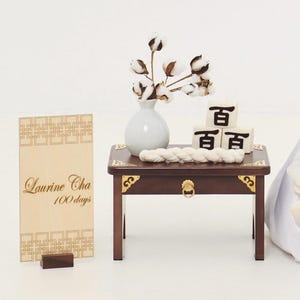 May include: A small, dark wooden table with gold accents holds a white vase with cotton stems, three stacked blocks with black characters, and a white rope. A wooden sign with the text "Laurine Cha 100 days" stands to the left.