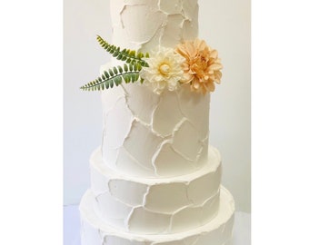 May include: A four-tier white wedding cake with a textured buttercream frosting. The top tier is decorated with a cluster of peach and white flowers and green foliage.