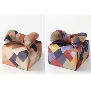 Puede incluir: Dos cajas de regalo envueltas en tela con patrones geométricos coloridos. Una caja presenta un lazo con tonos naranja, beige y marrón, mientras que la otra tiene un lazo con tonos morados, amarillos y rojos. Las cajas se presentan sobre un fondo blanco.