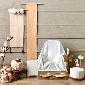 May include: A white, satin-covered chair with a wooden base sits in front of a light gray wood wall. Two beige fabric banners hang on the wall, one with white cotton balls and the other with a simple design. A white cake with a wooden base sits on a wooden table with two gold metal trays. A wooden candle holder with a gold butterfly design and a white candle sits on the table. A white flower with a wooden stick and a Korean character is placed on the cake.