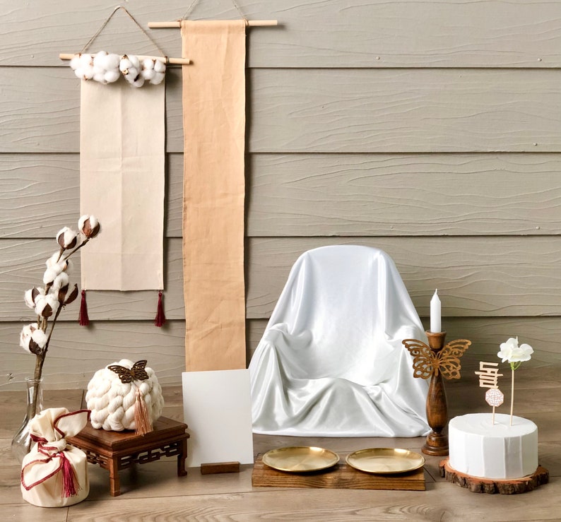 May include: A white, satin-covered chair with a wooden base sits in front of a light gray wood wall. Two beige fabric banners hang on the wall, one with white cotton balls and the other with a simple design. A white cake with a wooden base sits on a wooden table with two gold metal trays. A wooden candle holder with a gold butterfly design and a white candle sits on the table. A white flower with a wooden stick and a Korean character is placed on the cake.