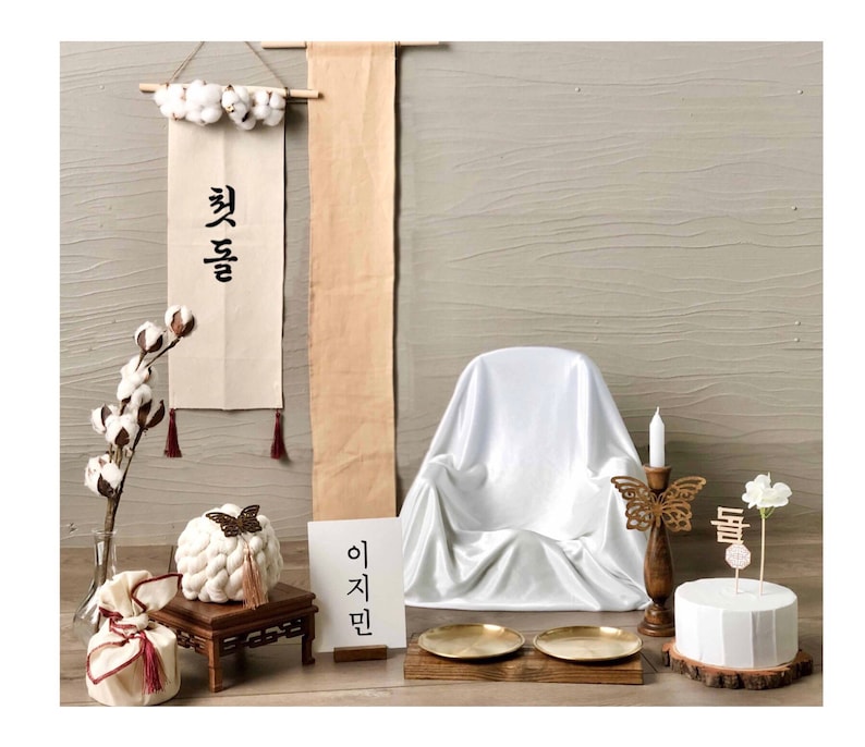 May include: A white backdrop with a white chair, a white cake with a wooden butterfly topper, a wooden candle holder with a white candle, and a white cotton flower arrangement. There are two wooden signs with Korean text, "첫돌" and "이지민".