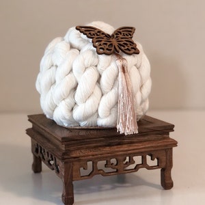 May include: A white crocheted ball with a wooden butterfly ornament and a pink tassel sits on a dark wood pedestal with intricate carvings.