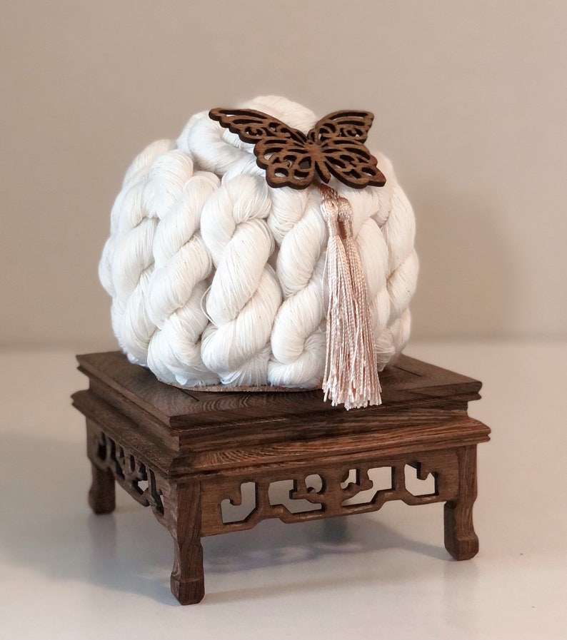 May include: A white crocheted ball with a wooden butterfly ornament and a pink tassel sits on a dark wood pedestal with intricate carvings.
