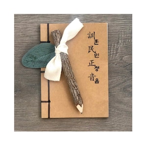 May include: A handmade journal with a brown cover, black stitching, and a wooden pencil tied with a white ribbon. A green leaf accent is tucked into the journal. Korean text is printed on the journal's cover.