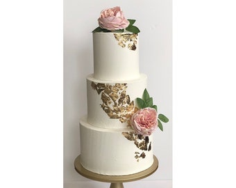 May include: A three-tiered white wedding cake decorated with gold leaf accents and pink roses.