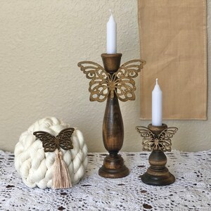 May include: Two wooden candle holders with butterfly designs. The larger holder has a white candle and the smaller holder has a white candle. Both holders are on a white crocheted surface.
