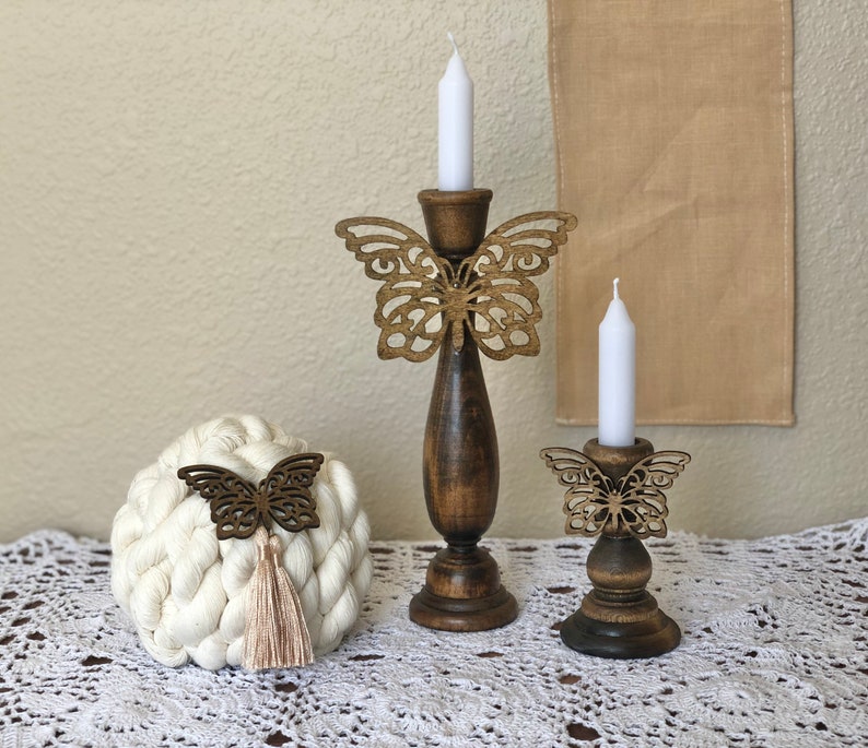 May include: Two wooden candle holders with butterfly designs. The larger holder has a white candle and the smaller holder has a white candle. Both holders are on a white crocheted surface.