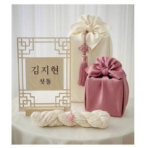 May include: An arrangement of Korean traditional gift wrapping, including a wooden sign with Korean text, two wrapped gifts in cream and pink, and a bundle of white yarn with a pink knot.