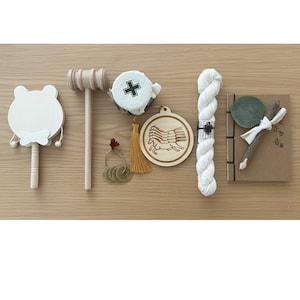 May include: An assortment of handcrafted items, including a white frog-shaped rattle, a wooden mallet, a round wooden pendant with a horse design, a bundle of white yarn, and a small notebook with a pen. The items are arranged on a light brown wooden surface.