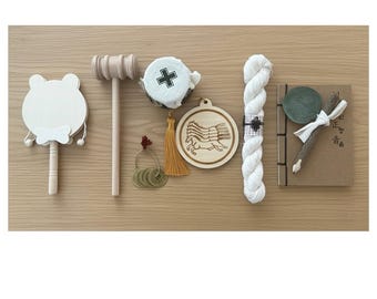 Peut inclure: Divers objets artisanaux, dont un hochet blanc en forme de grenouille, un maillet en bois, un pendentif rond en bois avec un motif de cheval, une pelote de fil blanc et un petit carnet avec un stylo. Les objets sont disposés sur une surface en bois brun clair.