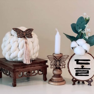 May include: A white crocheted ball with a wooden butterfly embellishment sits on a wooden stand. A wooden candle holder with a butterfly design holds a white candle. A wooden sign with the Korean word "Dul" carved into it sits on a small wooden stand. The sign is decorated with cotton balls and green leaves.