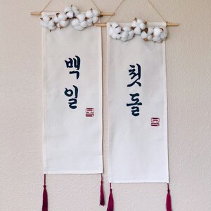 May include: Two white fabric banners with Korean calligraphy and red tassels hanging from a white wall with cotton flower garlands at the top.
