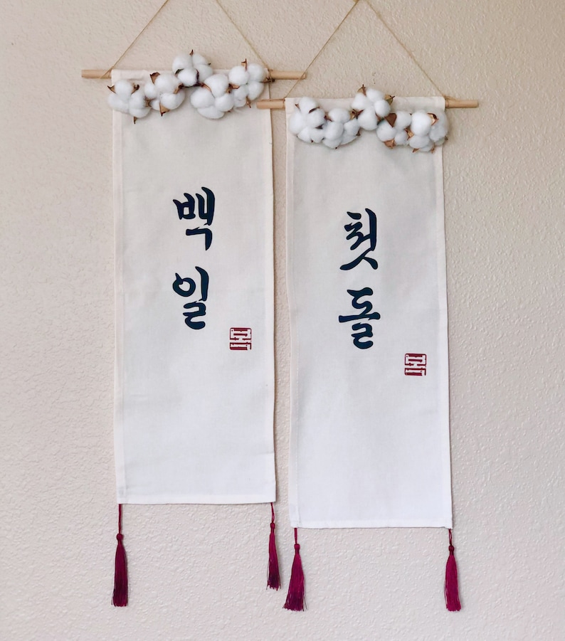 May include: Two white fabric banners with Korean calligraphy and red tassels hanging from a white wall with cotton flower garlands at the top.