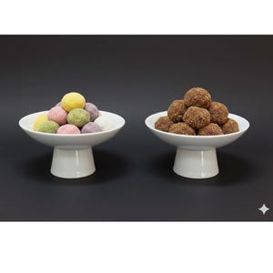 May include: Two white pedestal bowls on a black background. One bowl holds colorful, round treats in yellow, pink, green, purple, and white. The other bowl contains brown, spherical treats. The bowls have a simple, elegant design.