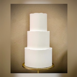 May include: A three-tiered white cake on a gold stand. The cake is plain and undecorated.