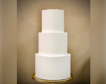 May include: A three-tiered white cake on a gold stand. The cake is plain and undecorated.