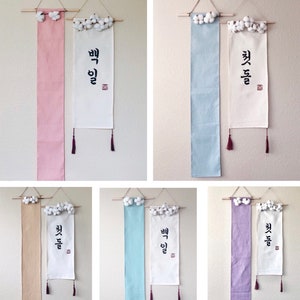 May include: Six white fabric banners with Korean calligraphy and cotton balls hanging from the top. The banners are in various colors: pink, light blue, beige, light green, and purple.