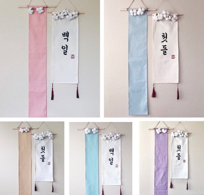 May include: Six white fabric banners with Korean calligraphy and cotton balls hanging from the top. The banners are in various colors: pink, light blue, beige, light green, and purple.