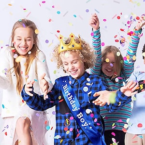 Puede incluir: Un grupo de ni&ntilde;os celebrando un cumplea&ntilde;os. Un ni&ntilde;o lleva una corona dorada y una banda azul que dice "Birthday Boy". Los ni&ntilde;os est&aacute;n rodeados de confeti de colores.