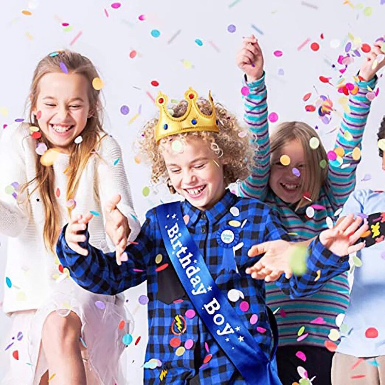 Puede incluir: Un grupo de ni&ntilde;os celebrando un cumplea&ntilde;os. Un ni&ntilde;o lleva una corona dorada y una banda azul que dice "Birthday Boy". Los ni&ntilde;os est&aacute;n rodeados de confeti de colores.