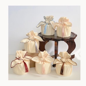May include: Six decorative gift boxes wrapped in cream-colored fabric with colorful tassels. The boxes are arranged on a wooden table.
