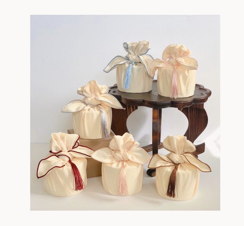 May include: Six decorative gift boxes wrapped in cream-colored fabric with colorful tassels. The boxes are arranged on a wooden table.