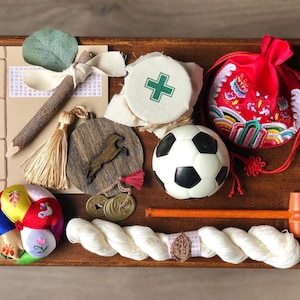 May include: A collection of decorative items on a wooden surface. Includes a soccer ball, a red embroidered pouch, a colorful fabric ball, a wooden hammer, a pencil with a leaf, and other small ornaments. The items are arranged on a dark brown wooden tray.