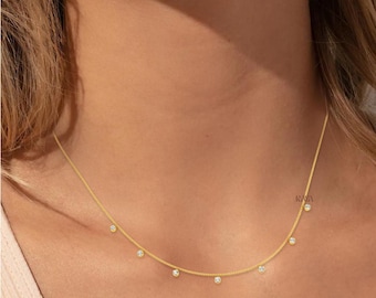 May include: A delicate gold chain necklace featuring seven evenly spaced, small round clear stones set in gold bezels. The necklace rests against the neck, showcasing a minimalist design with a thin, polished gold link chain and sparkling accents.