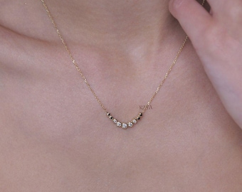 May include: A delicate gold necklace with a curved row of white and black gemstones. The necklace is worn on a person's neck, and a diamond ring is visible on their finger. The necklace has the word "KAYA" on it.