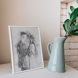 May include: A white picture frame with a black and white pencil drawing of a couple dancing. The frame is sitting on a white surface next to a light blue ceramic pitcher with a green plant.
