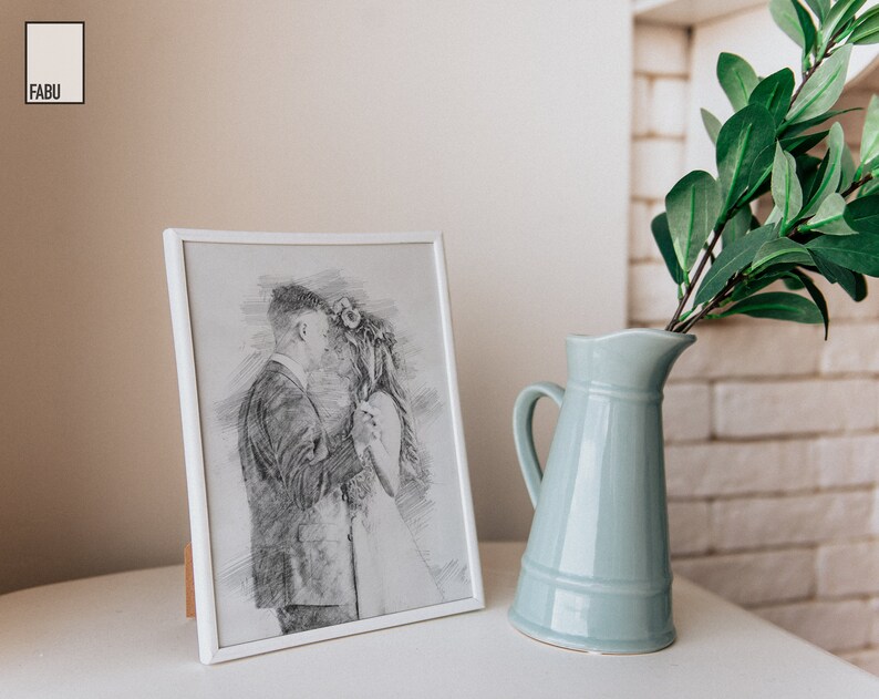 May include: A white picture frame with a black and white pencil drawing of a couple dancing. The frame is sitting on a white surface next to a light blue ceramic pitcher with a green plant.