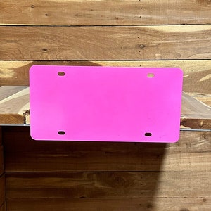 May include: A bright pink plastic license plate blank with four holes for mounting. The plate is hanging on a wooden wall.