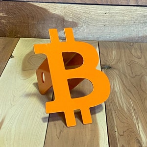 May include: Orange metal Bitcoin symbol hitch cover. The symbol is attached to a metal hitch receiver.