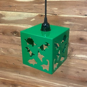 May include: A green metal pendant light fixture with a square shape and dinosaur cutouts. The light fixture is hanging from a black cord.