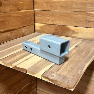 May include: A silver-colored metal trailer hitch receiver with a square opening and three circular holes. The hitch is designed to attach to a vehicle's frame for towing purposes. The hitch is shown on a wooden surface.
