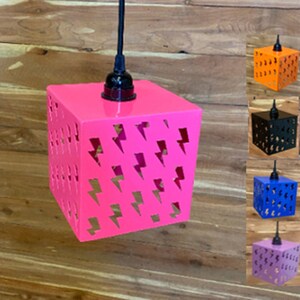 May include: A pink metal pendant light fixture with a black cord and a square shade. The shade has a lightning bolt pattern cut out of it. The light fixture is shown with other color options in the background.