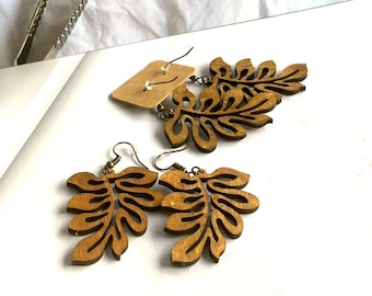 May include: Wooden leaf-shaped earrings with a natural wood finish. The earrings feature a detailed cutout design of leaves and are suspended from brass-colored ear wires. A small price tag is attached to one earring. The earrings are displayed on a white surface.