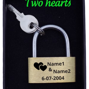May include: A brass padlock with a silver key, presented in a black box. The padlock features two black heart symbols and the text "Name1 & Name2" with the date "6-07-2004". The words "Two hearts" are written in green.