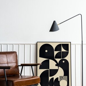 May include: A brown leather armchair with black metal legs sits in front of a black framed poster with a geometric design in black and beige. A black floor lamp with a cone-shaped shade is in the background.