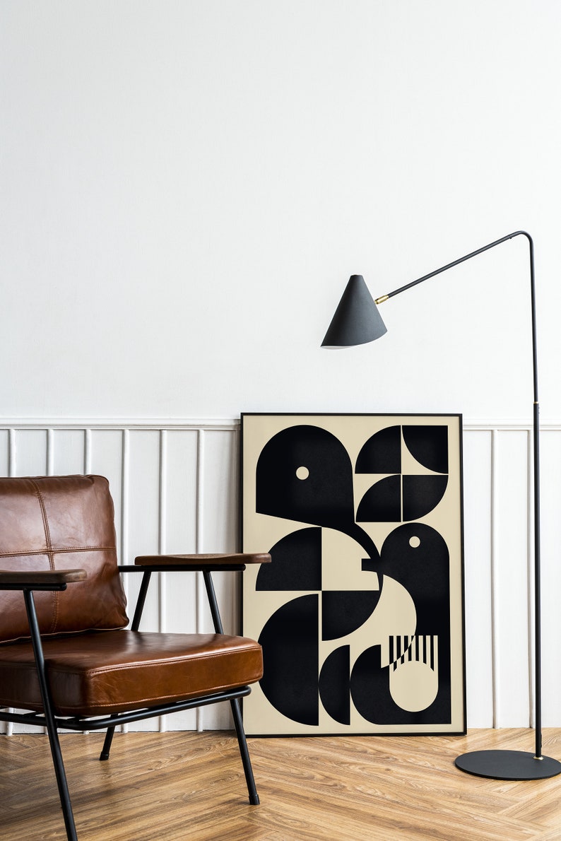 May include: A brown leather armchair with black metal legs sits in front of a black framed poster with a geometric design in black and beige. A black floor lamp with a cone-shaped shade is in the background.